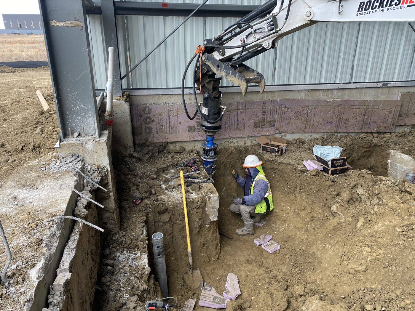Park Range crew member checking a helical pile as an excavator drives it beside a hangar foundation at Aero FNL