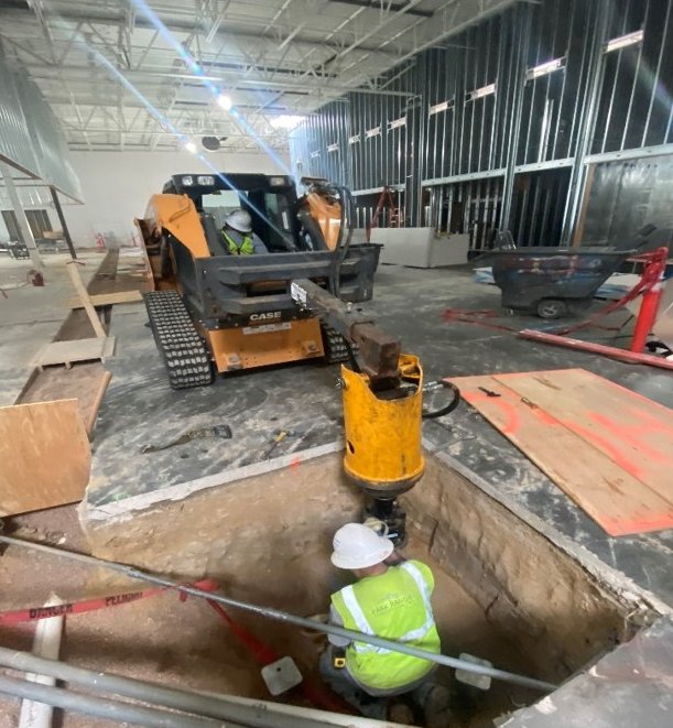 Park Range crew installing a 3-inch helical pile in an excavation inside the KSE Altitude Sports Radio building, with a compact track loader running the drive head
